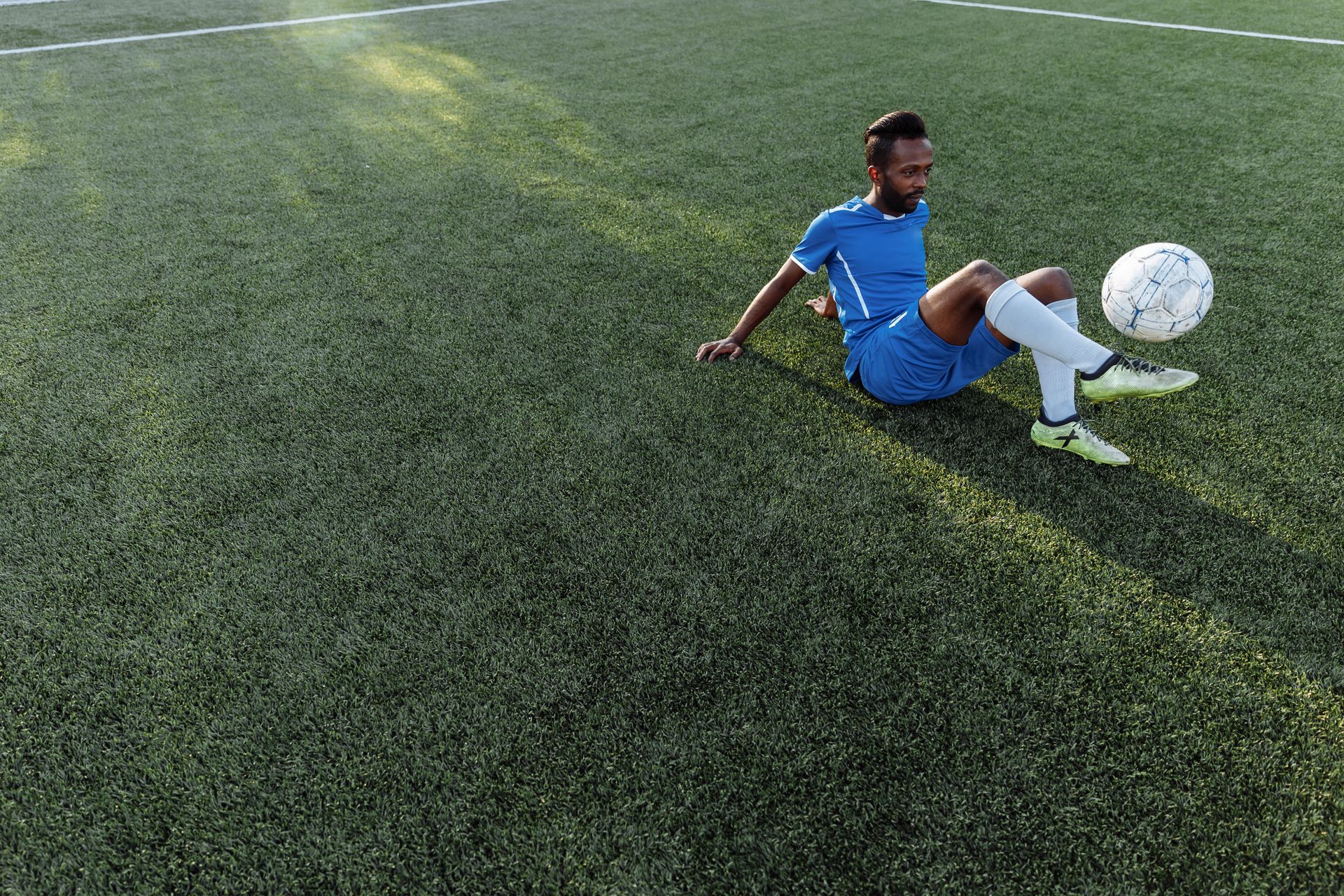 Man in Blue Shirt and Short Sitting While Kicking the Ball on Grass Field