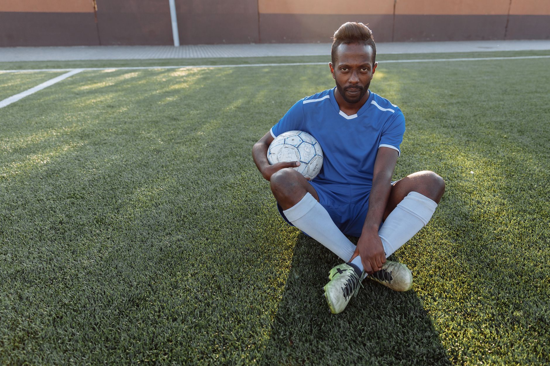 African man in blue jersey sitting on grass field holding a soccer ball.