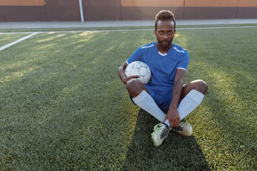 African man in blue jersey sitting on grass field holding a soccer ball.