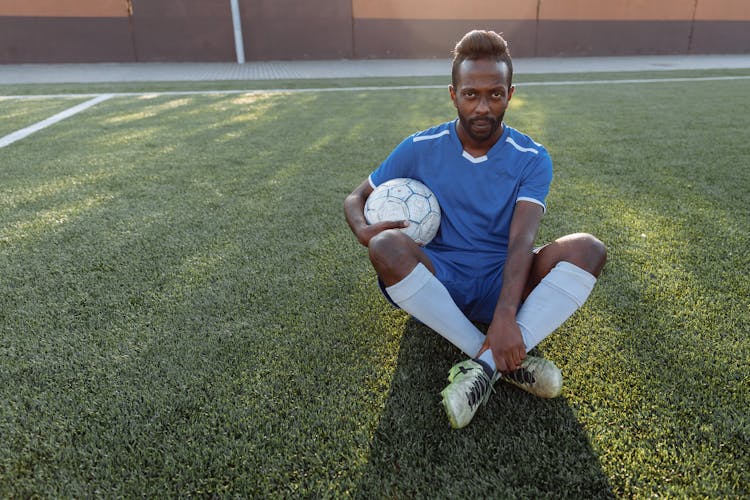 Man In Blue Jersey Holding Soccer Ball Sitting On Grass Field 