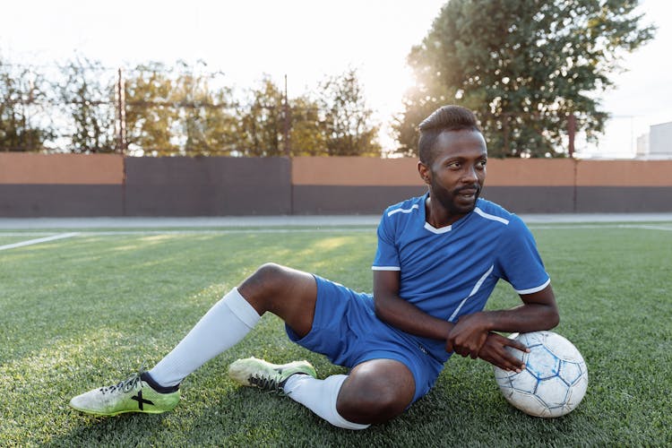 Man In Blue Football Jersey Sitting On Field Beside A Ball 