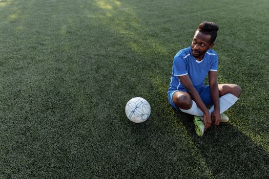A soccer player in a blue jersey sits on the grass field during the day.