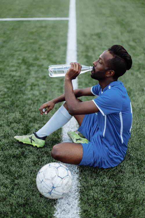 Football Player in Blue Jersey Sitting on Grass While Drinking Water 