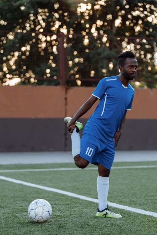 Free Soccer player in blue uniform stretching on a field with a ball nearby. Stock Photo