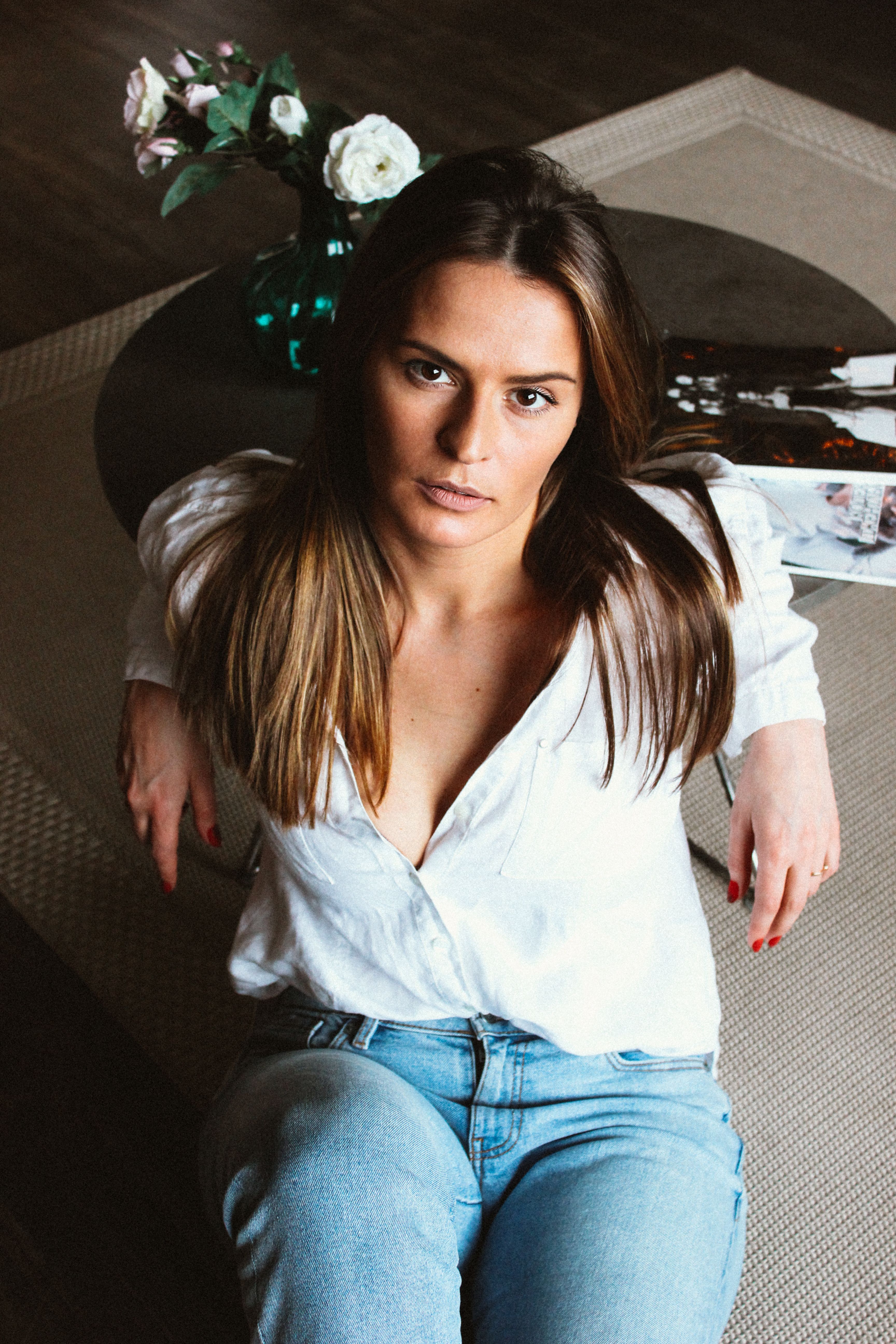 A woman in white blouse and jeans relaxing indoors, seated by a table.