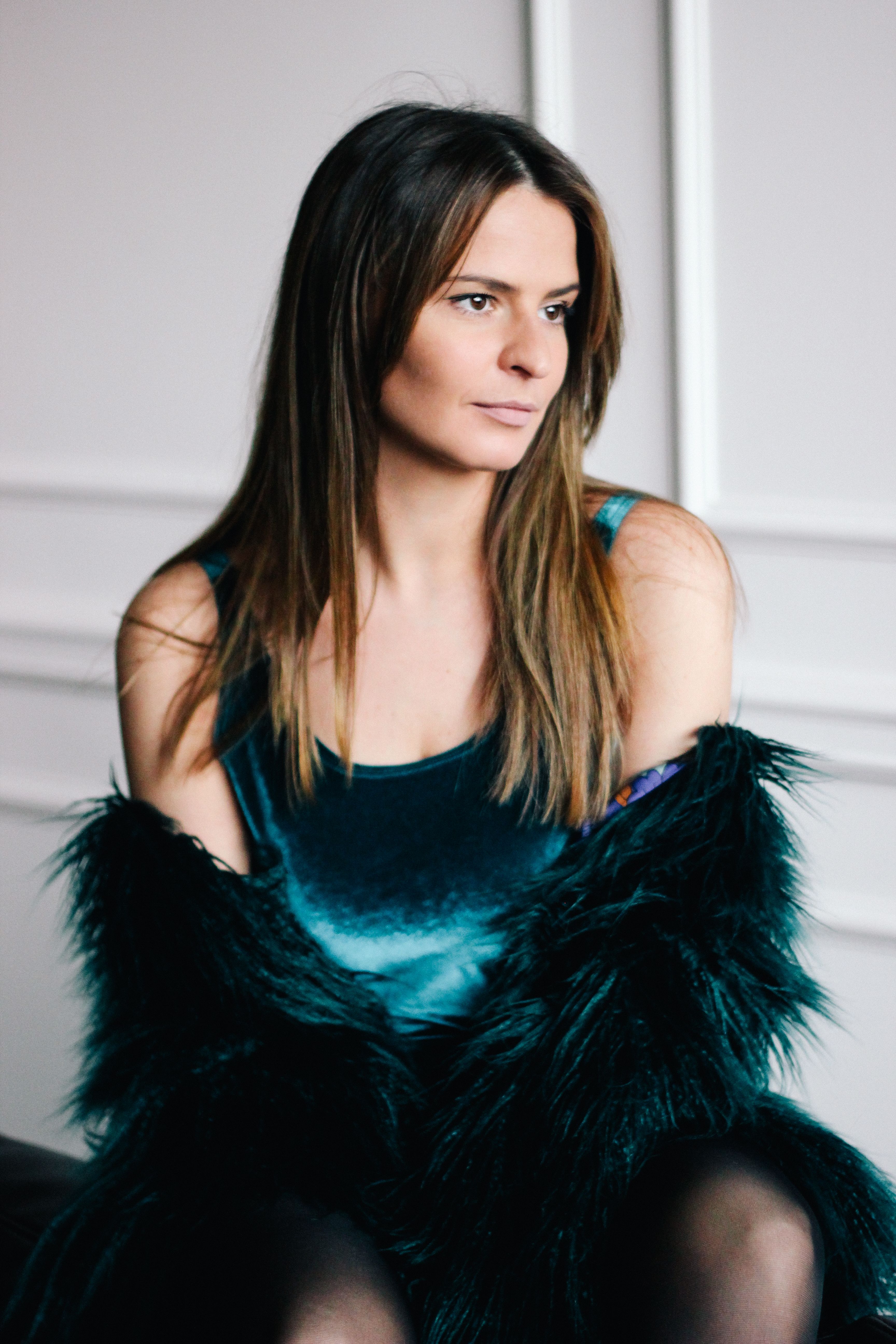 Portrait of a woman in a teal velvet dress and fur coat captured indoors against a bright background.