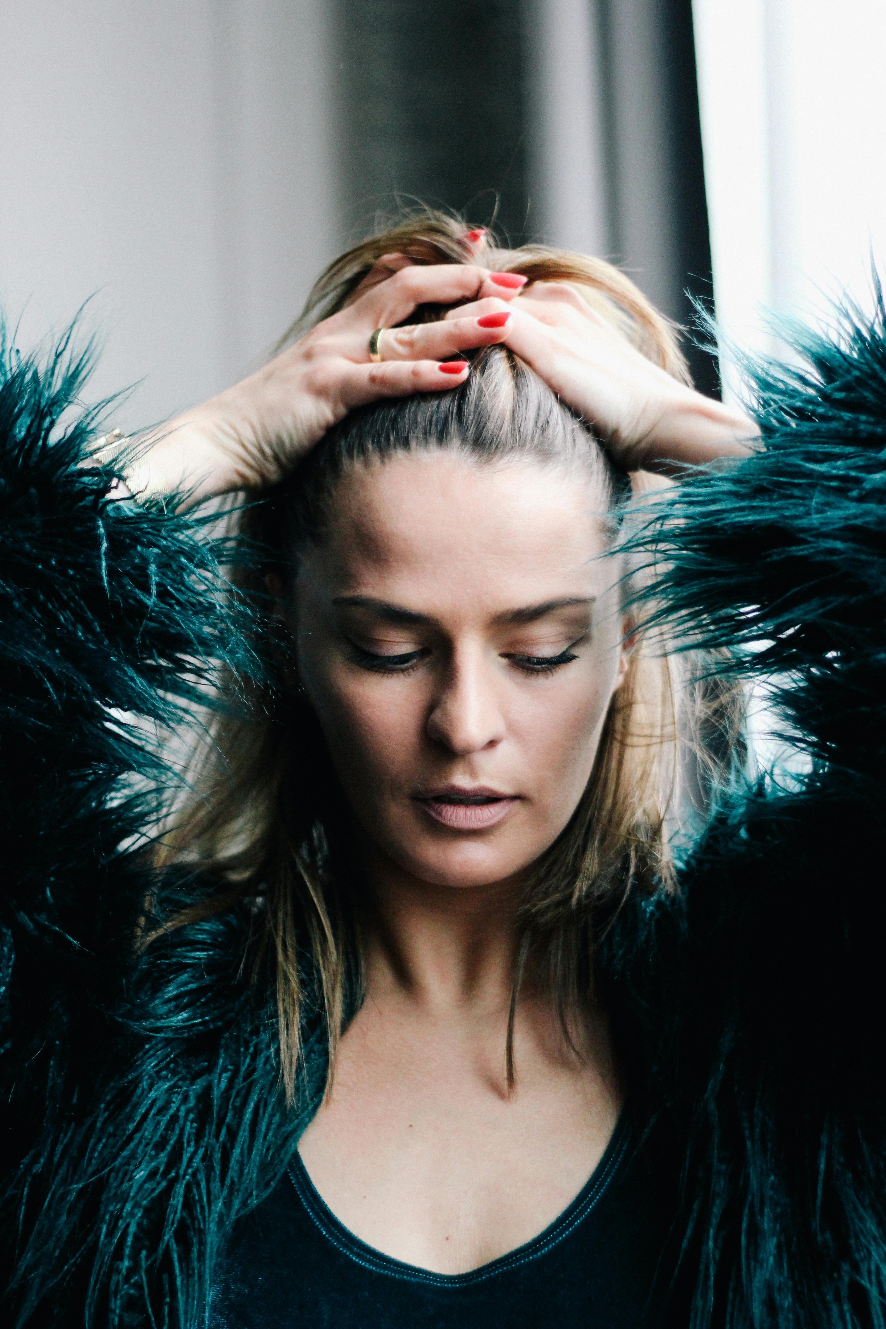 Close-up portrait of a contemplative woman in a dark faux fur coat by a window.