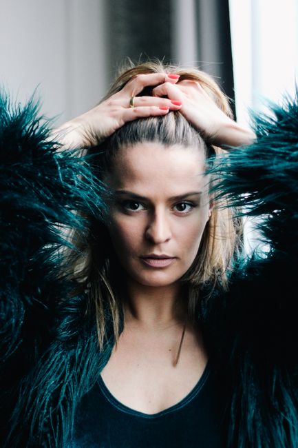 Close-up of a woman indoors, adjusting her hair while wearing a fluffy coat. Bright and stylish.