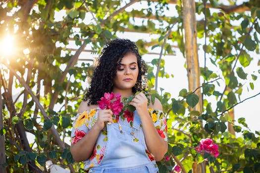 阳光明媚的花园里，一名穿着鲜艳服装的女子捧着粉红色的花朵，周围是郁郁葱葱的绿色植物