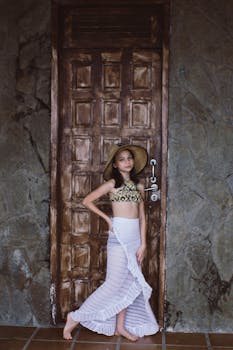 Young girl in a beach outfit and sun hat posing by a rustic wooden door.