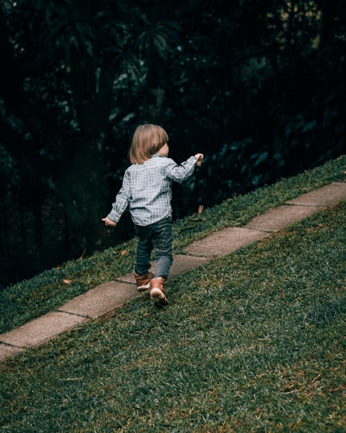 Back view full length cute little boy in stylish outfit strolling energetically on walkway in green summer park