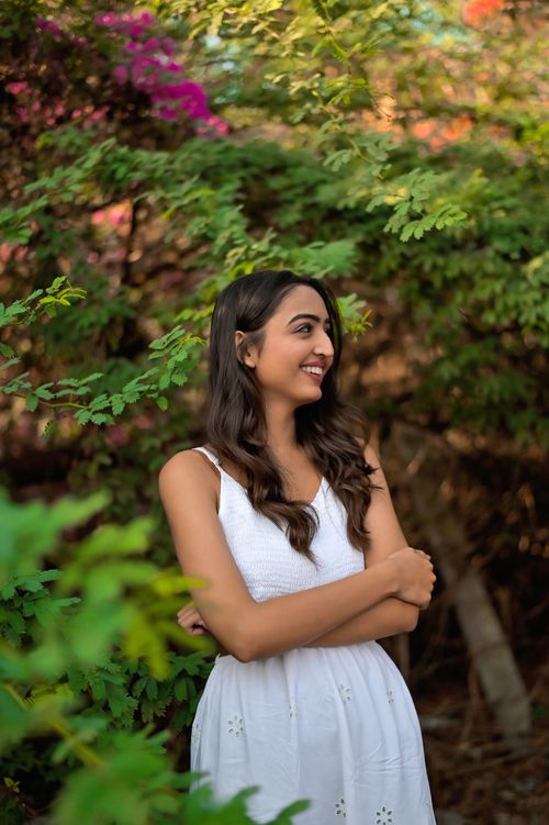 Free Smiling ethnic female with long wavy hair in white elegant dress standing with crossed arms on nature at daytime Stock Photo