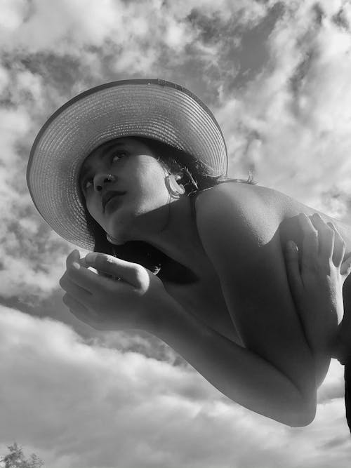 Free Black and white of young female with short hair in hat covering breast and standing against cloudy sky Stock Photo
