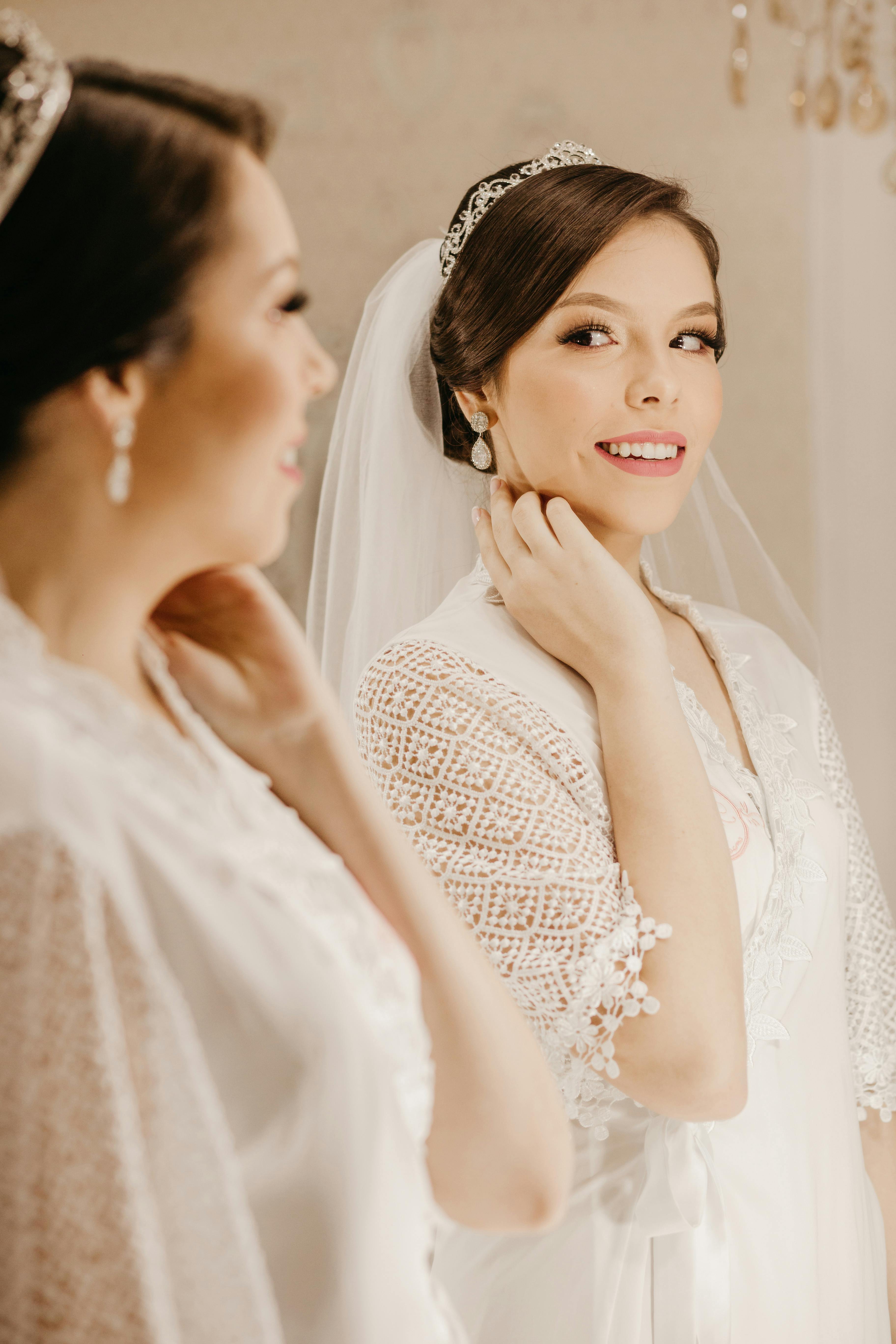 Cheerful young beautiful fiancee with hair tied up in white delicate dress reflecting in mirror while smiling and touching neck