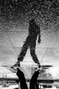Black and white of crop unrecognizable man in trendy wear with hat reflecting in glowing wet pavement on rainy day