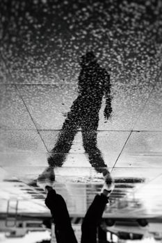 一名穿着时髦服装、戴着帽子的男子，在雨天发光的潮湿人行道上映出倒影，其身影与黑白相间的农作物背景融为一体