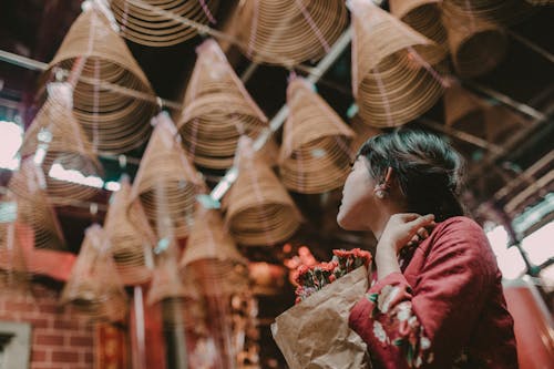 Free Low angle side view of unrecognizable Asian pilgrim with flower bouquet contemplating incense coils in Buddhist church Stock Photo