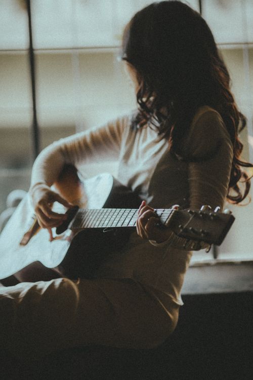 Free Anonymous female musician with hairstyle playing guitar while looking out window in house Stock Photo