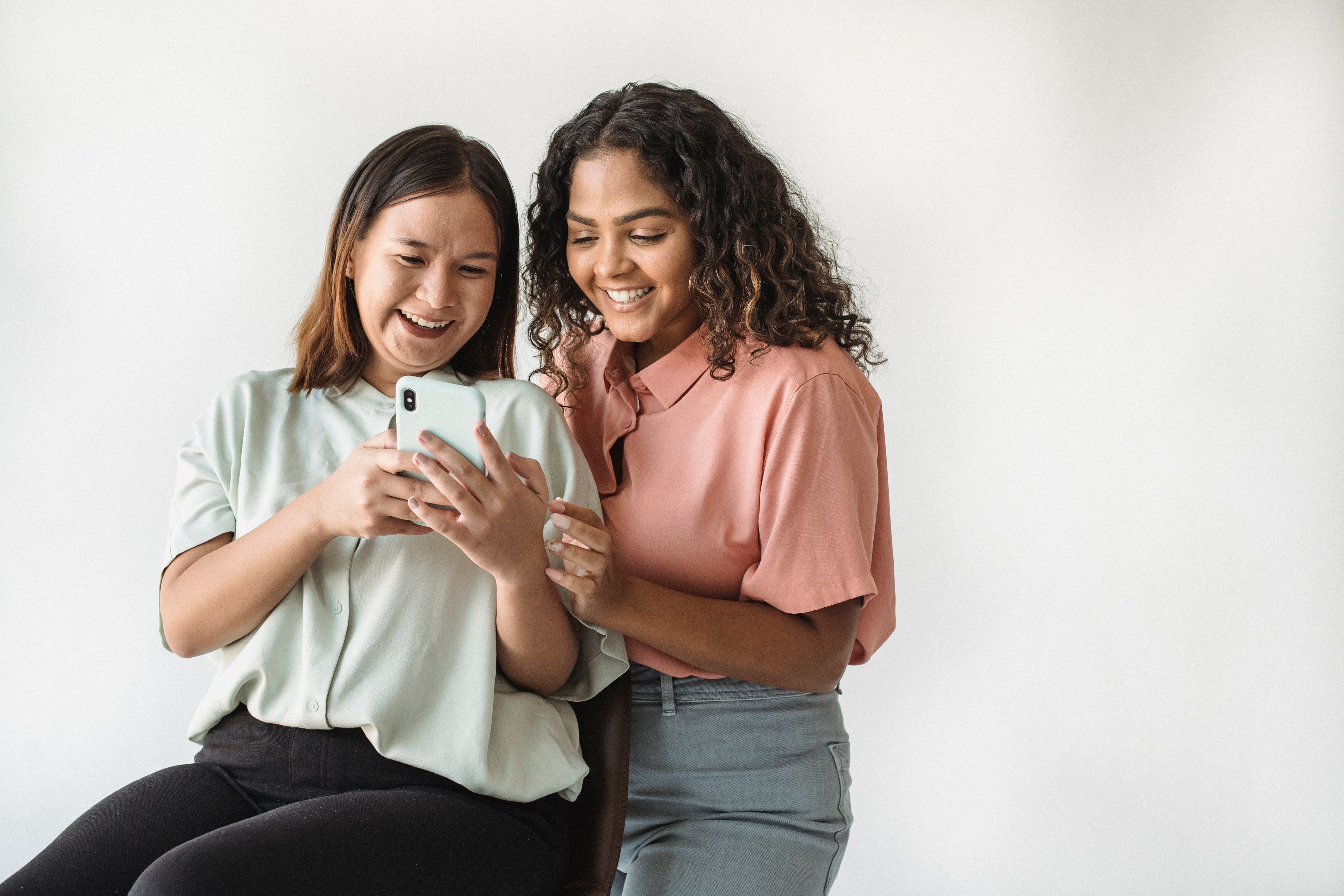 Two women smiling and looking at a smartphone together in a casual indoor setting.