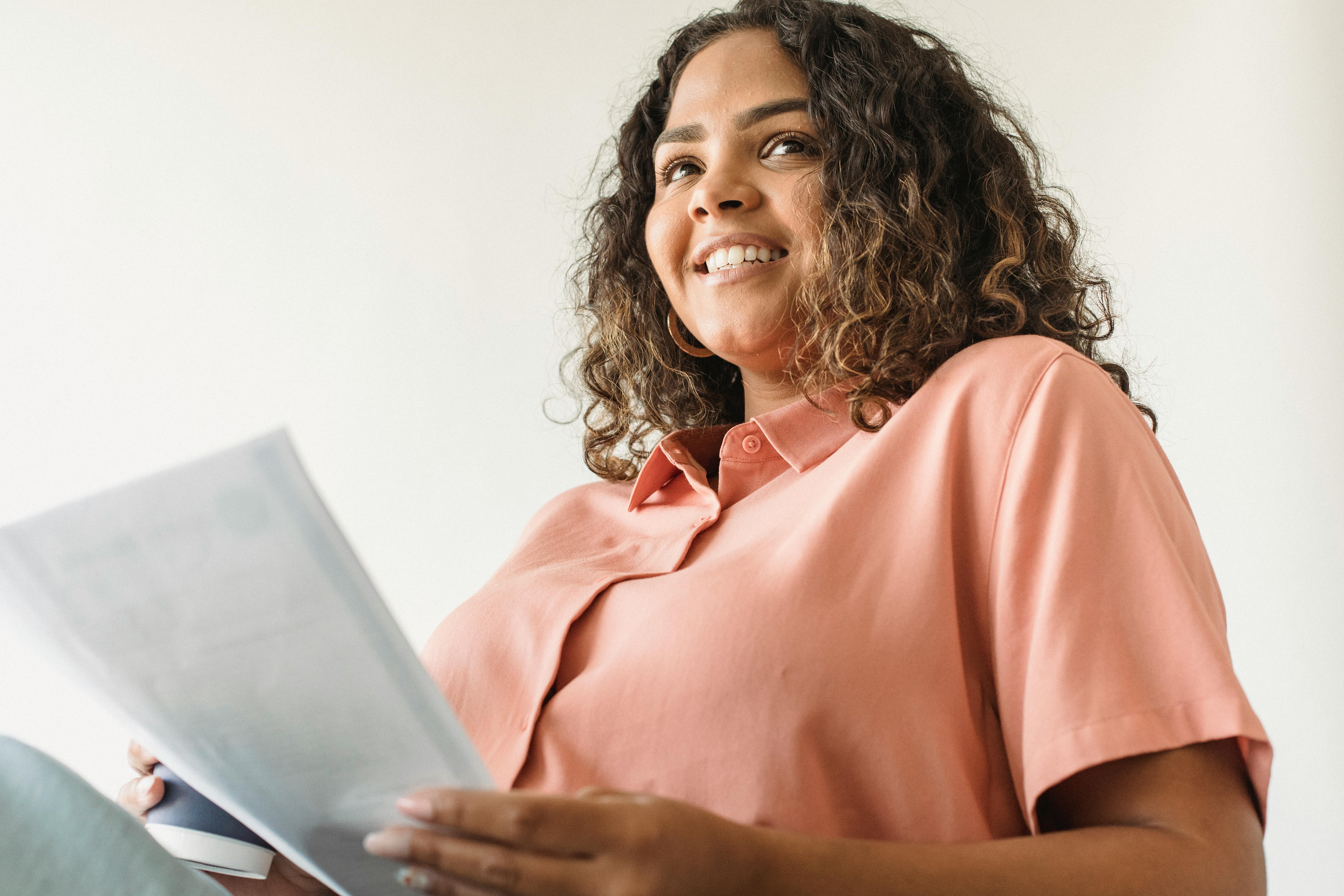 一位自信的年轻女子穿着粉色衬衫，在室内拿着文件微笑