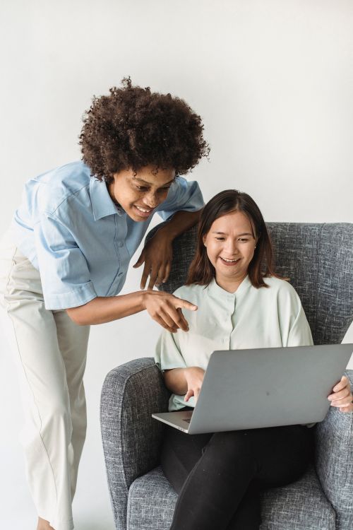 Free Two women collaborating on a project, one sitting and one standing, sharing a laptop and smiling. Stock Photo