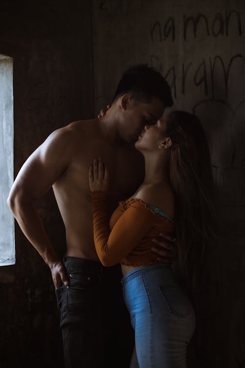 Side view of romantic couple embracing and kissing gently while standing near wall with lettering in dark abandoned building inside
