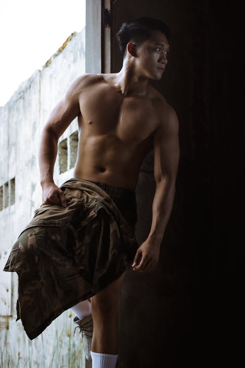 Muscular male with naked torso looking away while standing in doorway of shabby building with cape in hand in daylight