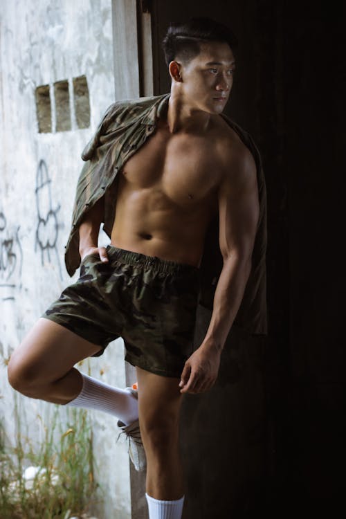 Serious male with naked torso wearing shorts leaning on shabby wall with obscene drawing while standing with hand in pocket and shirt on shoulder and looking away