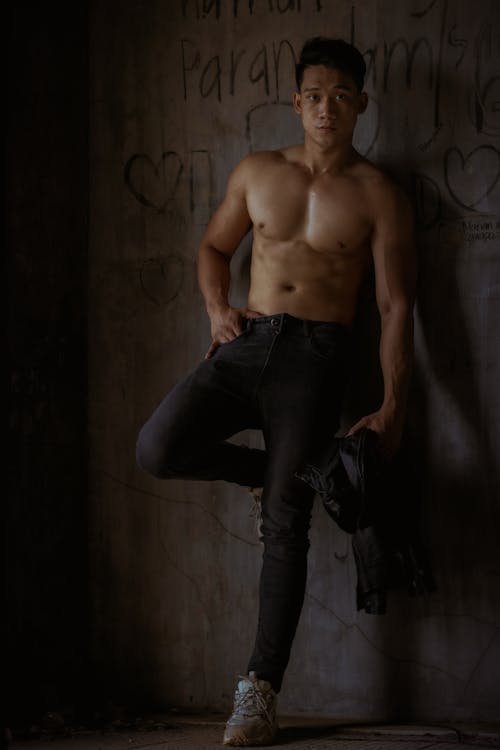 Full body of masculine male with naked torso looking at camera while standing near shabby wall with hand on hip and leg bent