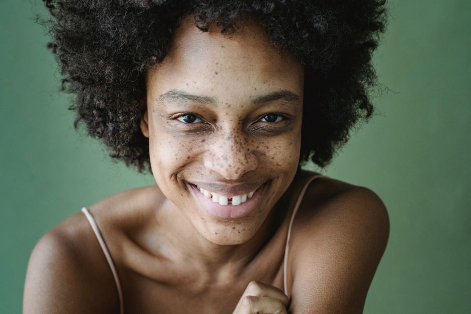 Portrait of a smiling woman with an afro hairstyle and freckles against a green background.