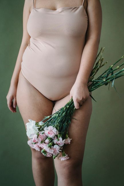 A woman stands holding a bouquet of flowers in a beige bodysuit against a green background, showcasing elegance and natural beauty.