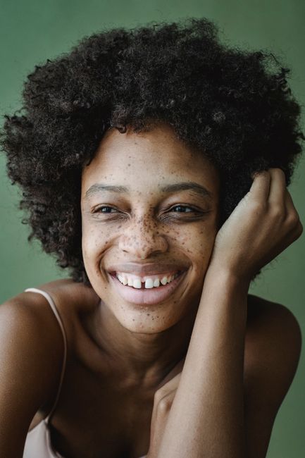 Close-up portrait of a smiling woman with natural curly hair and freckles against a green background.