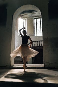 Silhouette of a ballerina in a tutu posing gracefully by large windows in an elegant vintage building.