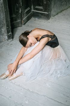 一位身着黑色上衣和白色芭蕾舞短裙的优雅芭蕾舞演员，正优雅地坐在室内的木地板上