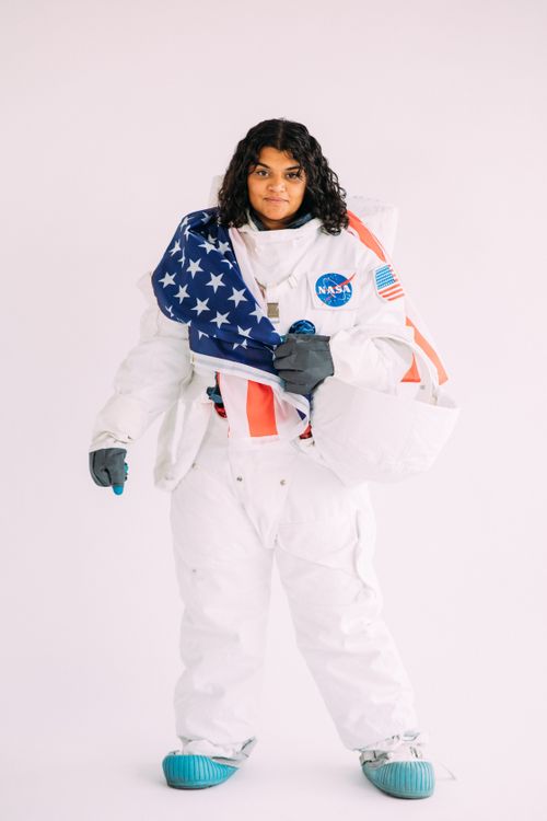 Free Portrait of a woman in a NASA astronaut suit holding an American flag against a plain backdrop. Stock Photo
