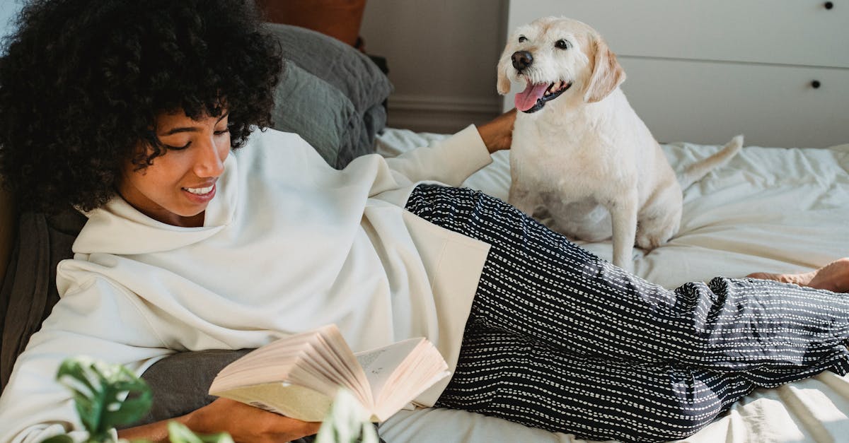 Smiling young black woman caressing dog while resting on bed and reading book