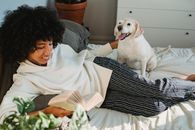 Smiling young black woman caressing dog while resting on bed and reading book