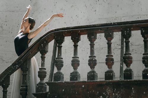 Free A graceful ballerina poses elegantly with hands raised beside a vintage staircase. Stock Photo