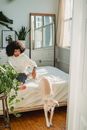 African American barefoot female sitting on bed with dog jumping on floor in comfortable room