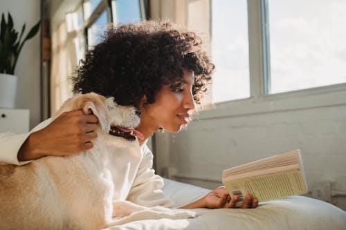 Black woman caressing dog while reading book on bed