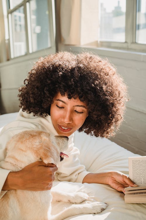 Black woman hugging dog while reading book in bedroom