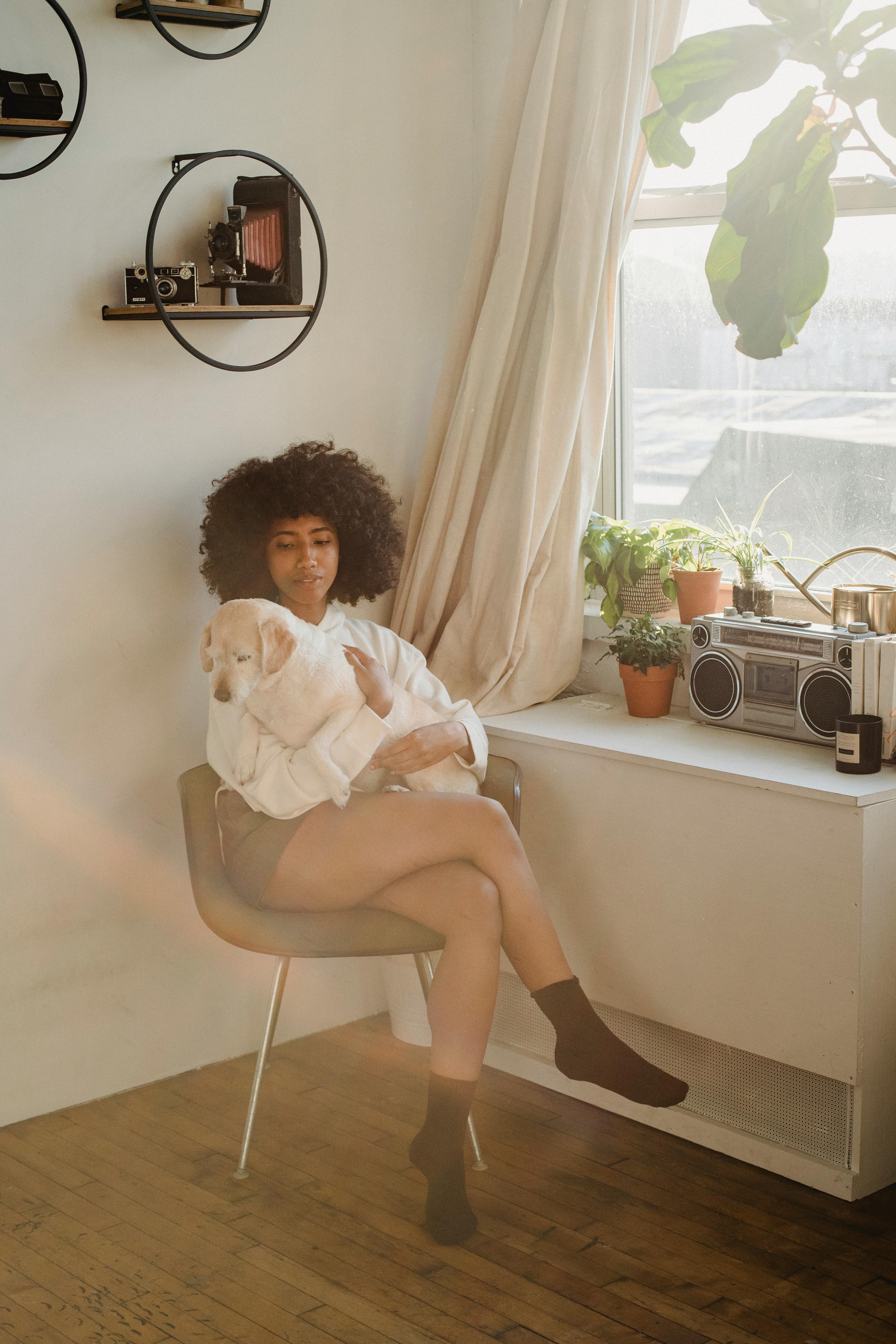 一位年轻的非裔美国女性身穿休闲服，坐在窗边，双腿交叉，怀里抱着一只狗