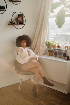 一位年轻的非裔美国女性身穿休闲服，坐在窗边，双腿交叉，怀里抱着一只狗