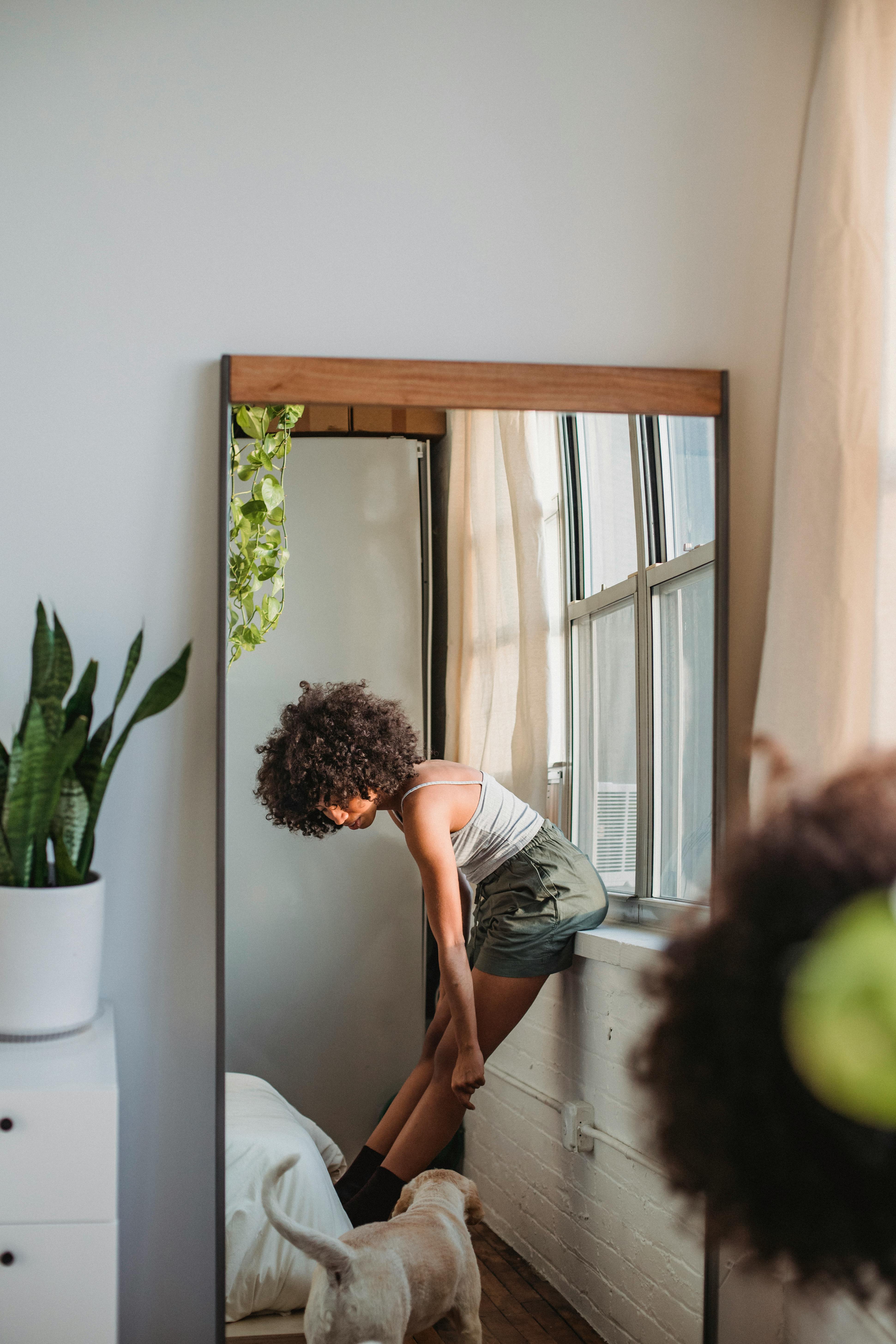 Side view of young African American female in casual wear with curly hair standing near window with dog and reflected in mirror