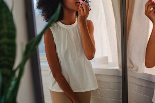 Crop black woman applying lip gloss against mirror