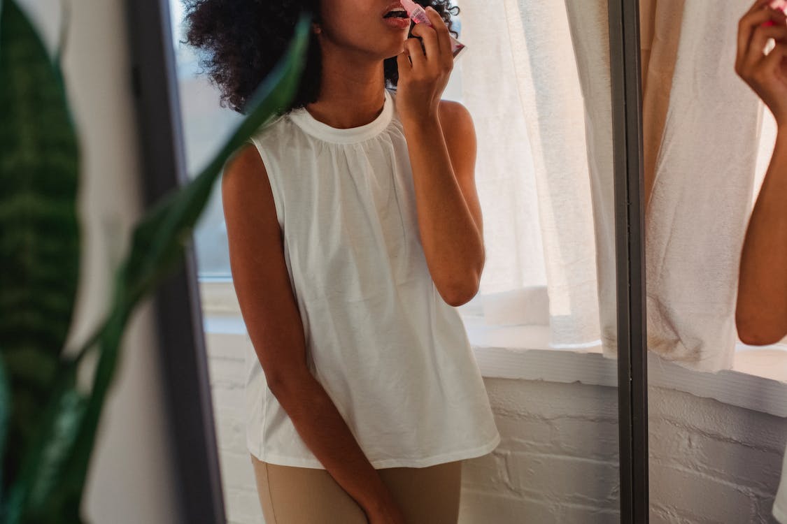 Crop black woman applying lip gloss against mirror