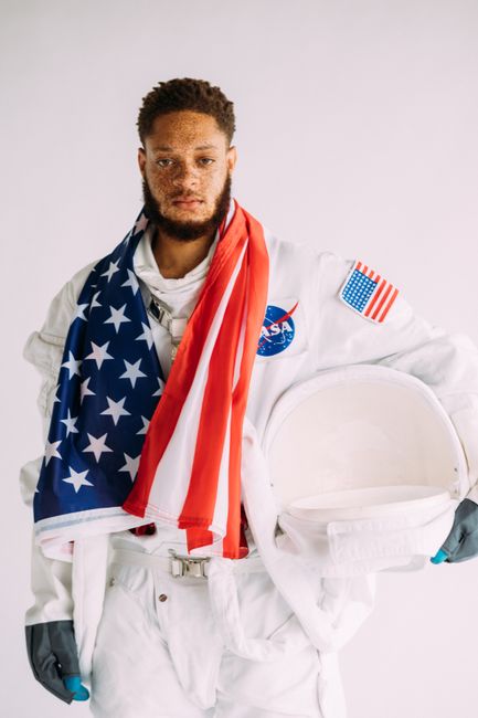 Astronaut portrait in space suit holding helmet, draped in American flag. White background.