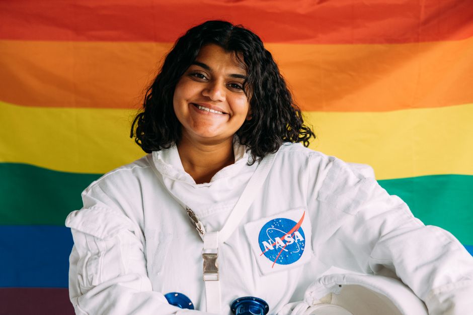 Smiling woman in NASA space suit posing with a pride flag background, celebrating diversity and space exploration.