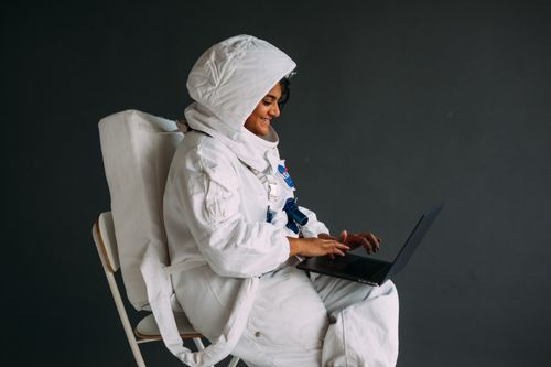 Free Smiling astronaut in white space suit sits indoors using a laptop, showcasing technology and exploration. Stock Photo