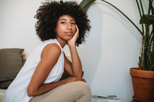 Free Side view of peaceful African American female with curly hair touching face while resting at home with potted plant Stock Photo